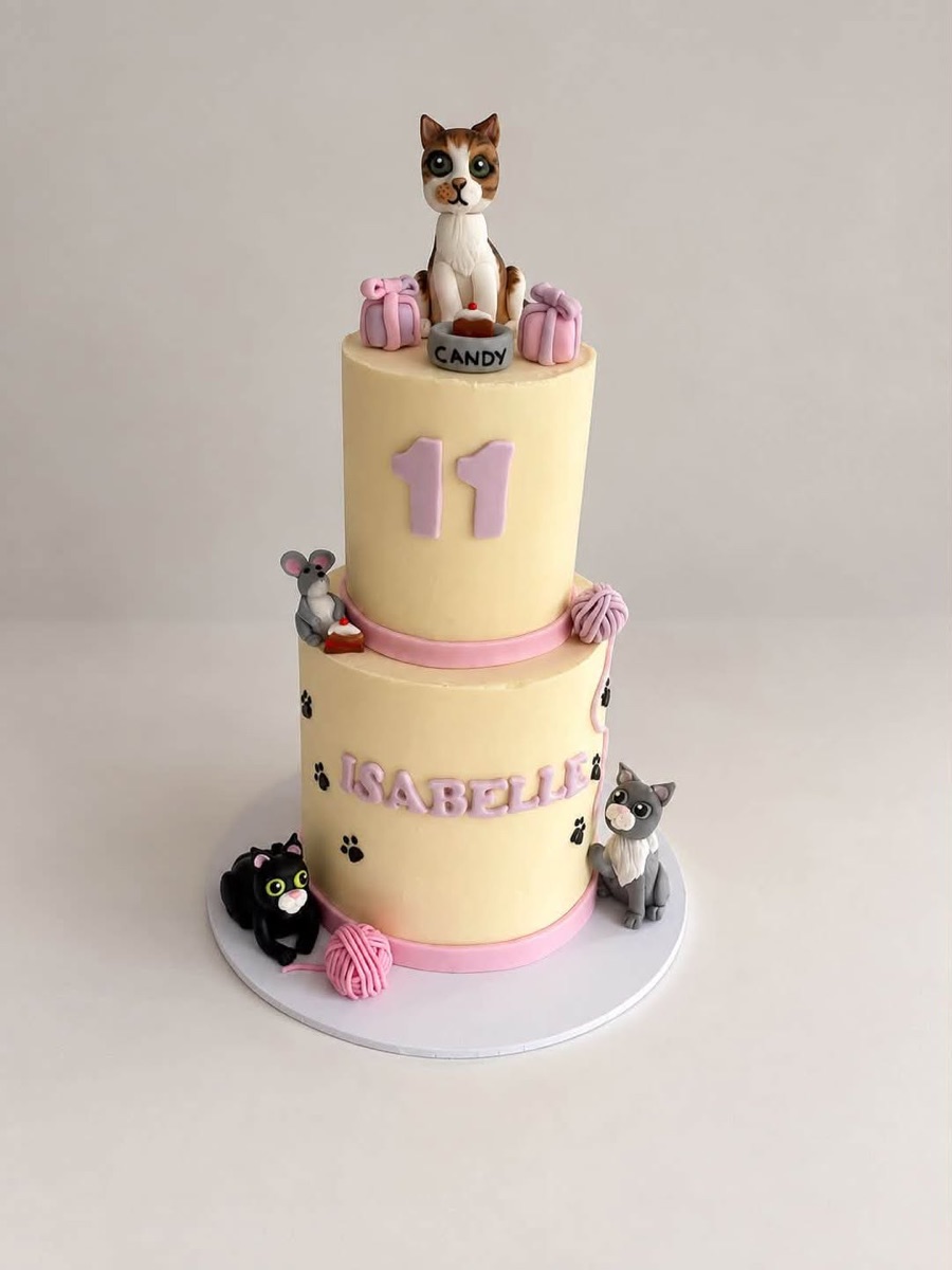Two tier buttercream cake with modelled cats, yarn balls and paw prints, topped with a tabby cat beside a cake bowl