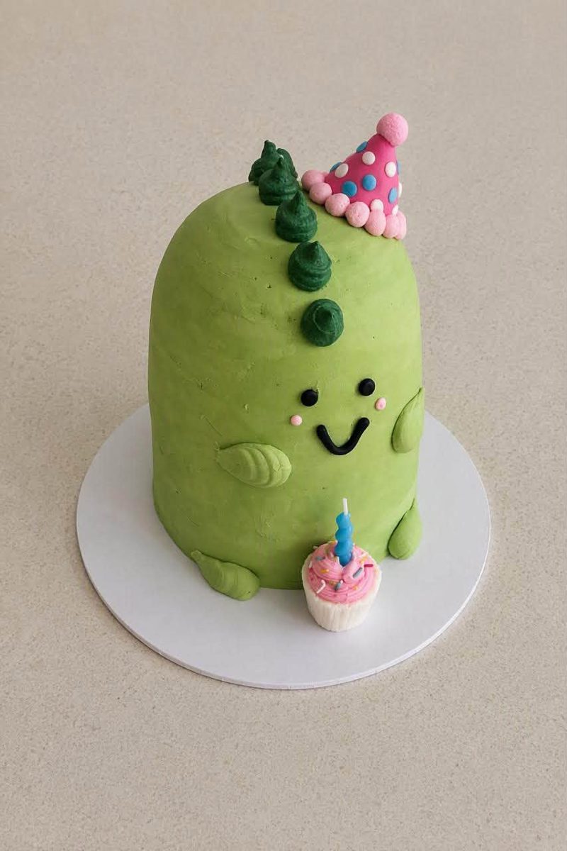 Green dinosaur cake wearing a party hat, with a mini cupcake