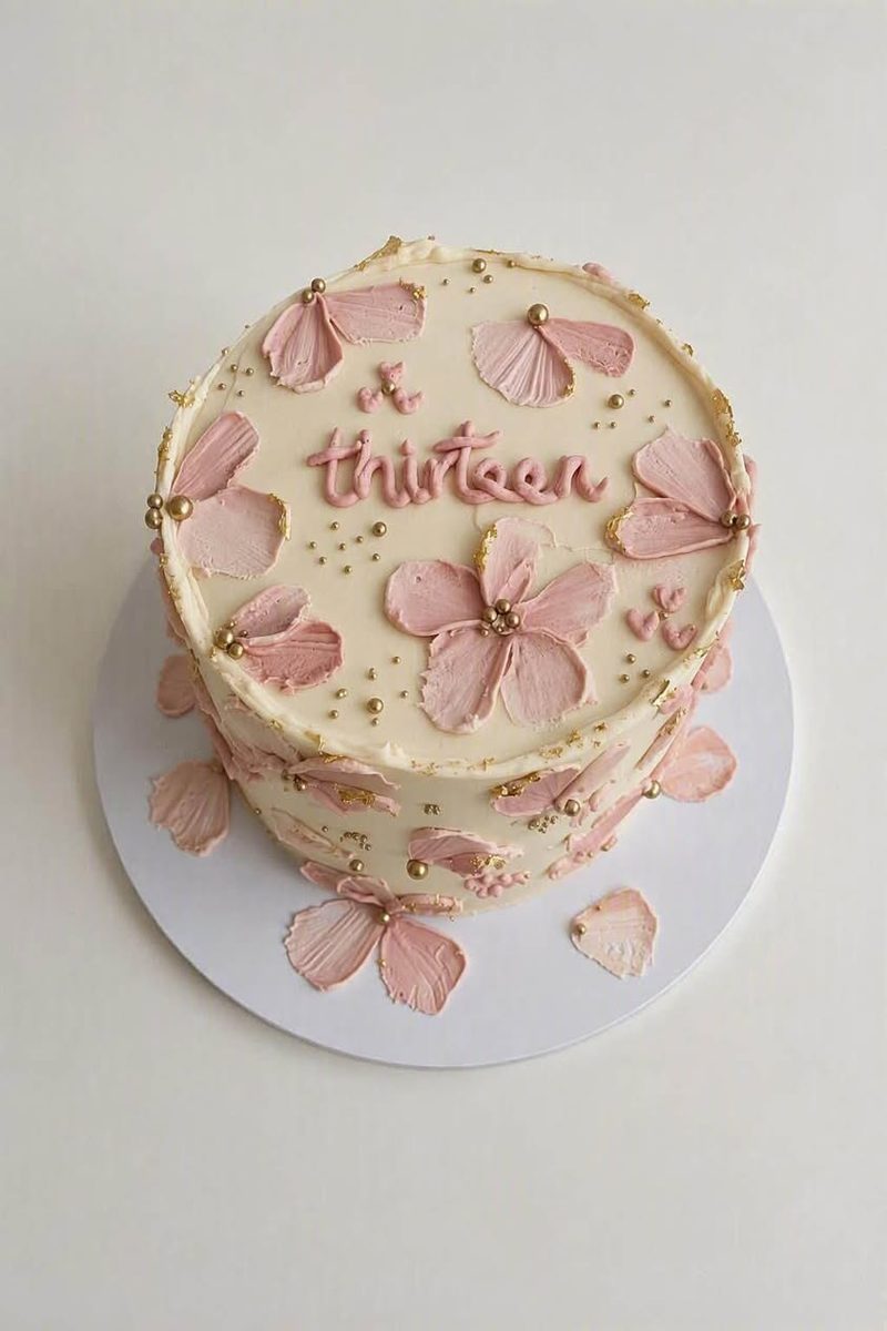 Cream buttercream cake with palette-knife painted pink flowers, gold accents and thirteen piped on top
